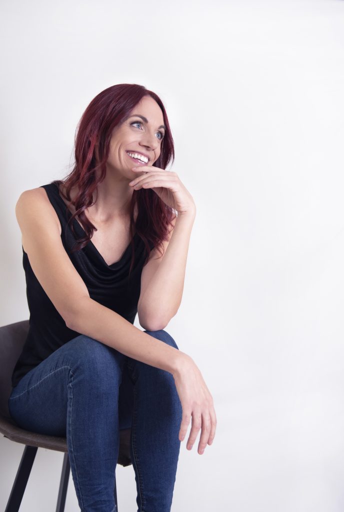 Femme souriante aux cheveux rouges, assise sur une chaise, posant dans un décor épuré, illustrant la bienveillance et l’authenticité.